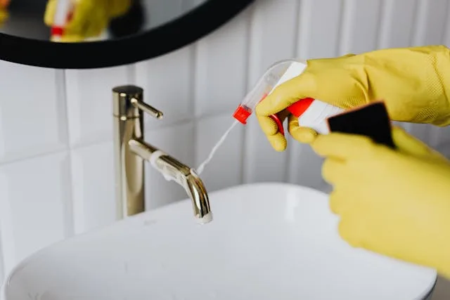 A close-up of a person wearing yellow gloves, spraying a cleaning solution onto a modern gold-colored bathroom faucet over a white sink.