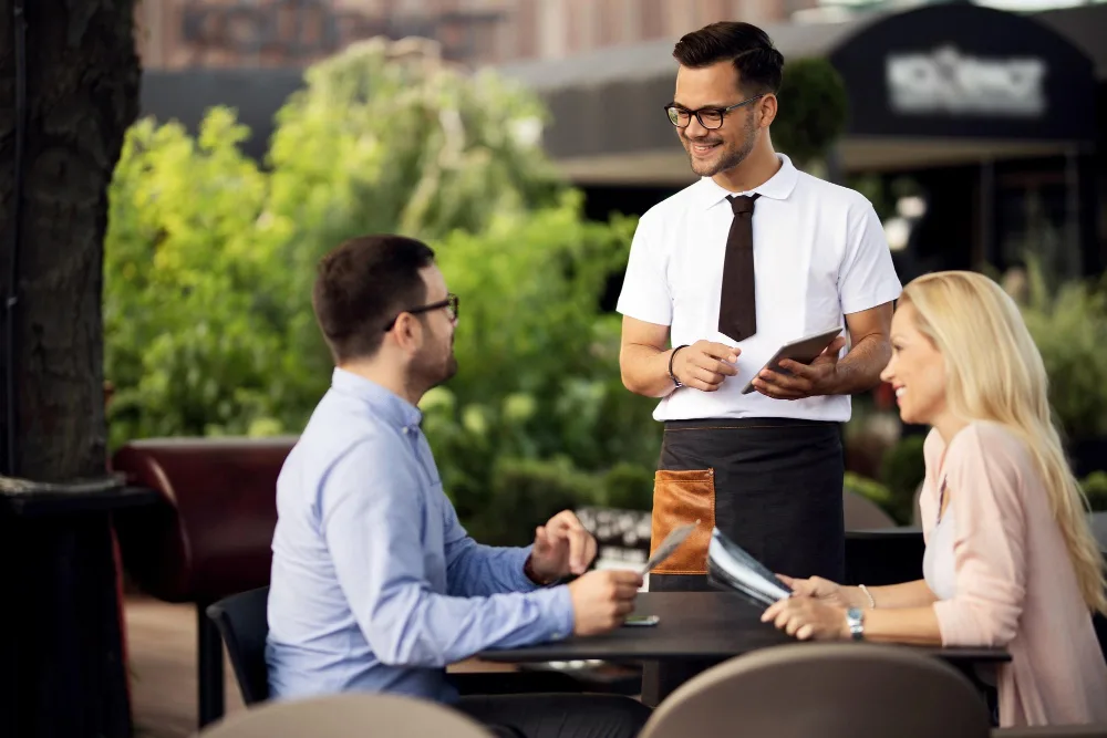 A smiling waiter in a white shirt, black tie, and apron takes an order from a man and a woman seated at an outdoor restaurant table.