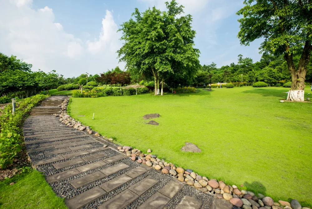 A well-maintained green park with a winding stone pathway bordered by decorative pebbles and lush green grass and trees under a partly cloudy sky.