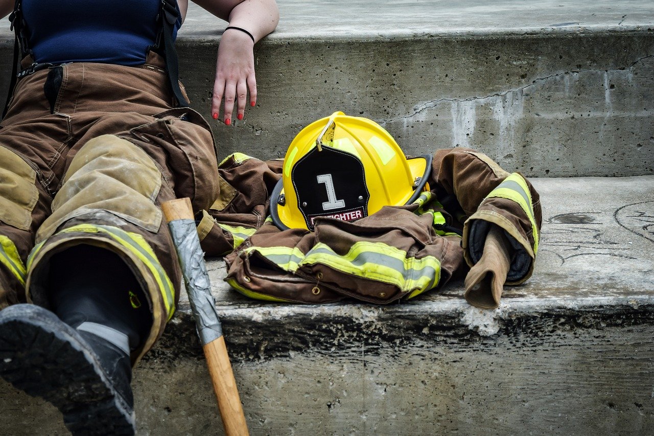 A firefighter in brown and yellow turnout gear rests on concrete steps next to their yellow helmet and jacket, with a tool nearby.