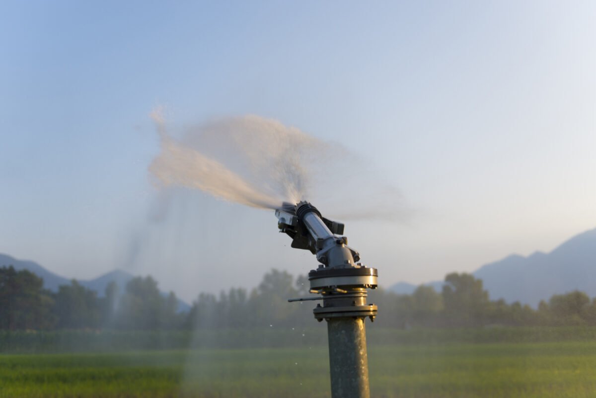 A large automatic irrigation sprinkler sprays water across a green field under a clear sky, with distant mountains and trees.